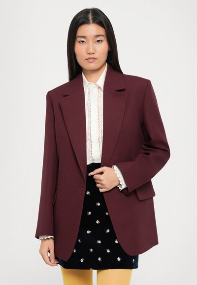 Burgundy blazer with a structured design, sharp lapels, and two front pockets, worn over a white shirt with lace detailing.