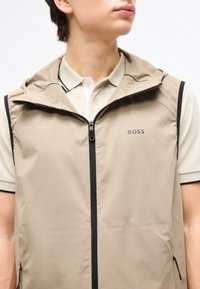 Νέος άνθρωπος που φοράει μπεζ αμάνικο γιλέκο με φερμουάρ και κουκούλα πάνω από κρεμ κοντομάνικο πόλο με μαύρες λεπτομέρειες.