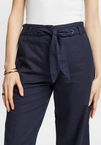 Mörkblå byxor med knytbart midjeband, bälteshållare och uppvikta fållar. Tyget har en mjuk struktur och en avslappnad passform.