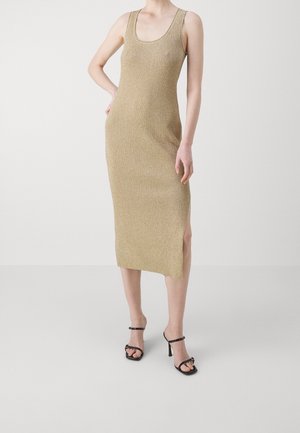 Kvinde iført ærmeløs midi-kjole i guld med sideslids og sorte åbne høje hæle, stående mod en ensfarvet baggrund.