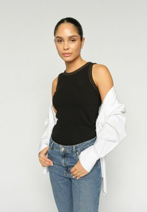 Kobieta z zaczesanymi do tyłu włosami, ubrana w czarny top bez rękawów, niebieskie dżinsy i białą koszulę zarzuconą na ramiona na jednolitym tle.