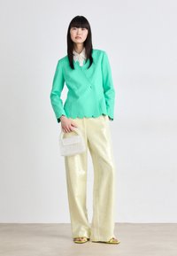 Mintgroene scalloped jas, crèmekleurige blouse, glanzende gele wijde broek, bewerkte witte handtas en gele sandalen met een eenvoudig ontwerp.