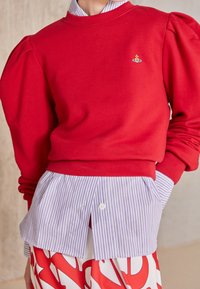 Sudadera roja con mangas abullonadas y cuello redondo, superpuesta sobre una camisa de rayas azules y blancas con cuello. Presenta un pequeño diseño bordado.