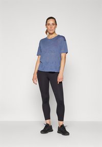 Kék sportpóló rövid ujjakkal, enyhén laza fazonban; fekete leggings és fekete sportcipő kombinálva. Simás textúra.