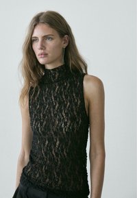 Zwarte kanten top met hoge hals en mouwloos ontwerp. Voorzien van ingewikkelde bloemmotieven en een doorschijnende textuur, gecombineerd met donkere broek.