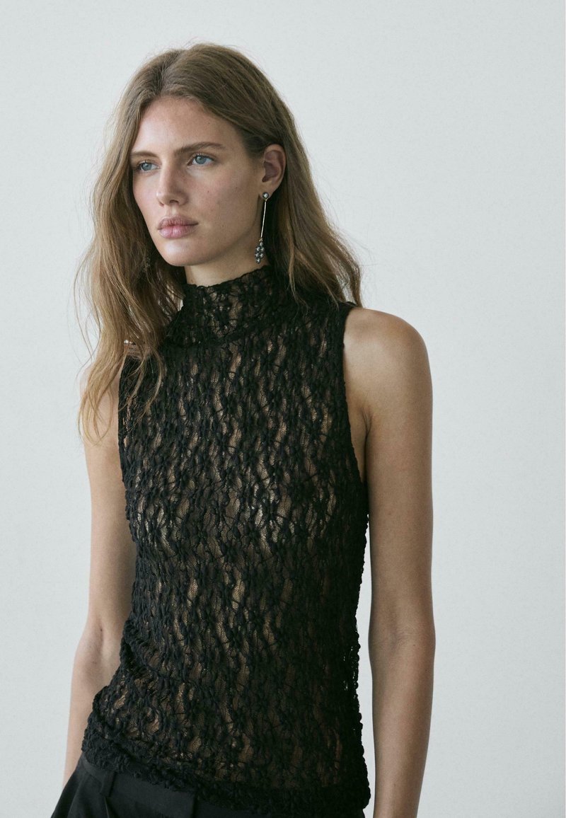 Zwarte kanten top met hoge hals en mouwloos ontwerp. Voorzien van ingewikkelde bloemmotieven en een doorschijnende textuur, gecombineerd met donkere broek.