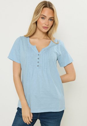 Młoda kobieta o blond włosach, ubrana w jasnoniebieską bluzkę z krótkim rękawem, zapinaną na guziki i ozdobioną pionowymi plisami, w połączeniu z ciemnoniebieskimi dżinsami.