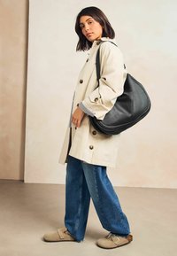 Beige trenchcoat med store knapper, sort læder skuldertaske, blå vide jeans og brune sandaler på en neutral baggrund.