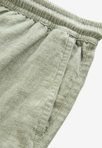 Gros plan sur un pantalon à taille élastique gris clair avec un tissu texturé et une couture de poche latérale verticale.