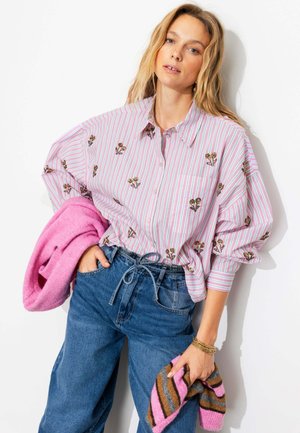 Femme portant une chemise rayée rose et blanche avec broderies florales, jean bleu taille haute, tenant une veste rose et une écharpe rayée.