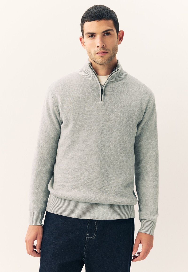 Ung mand iført en lys grå pullover med halv lynlås og mørke bukser, der står mod en ensfarvet baggrund.