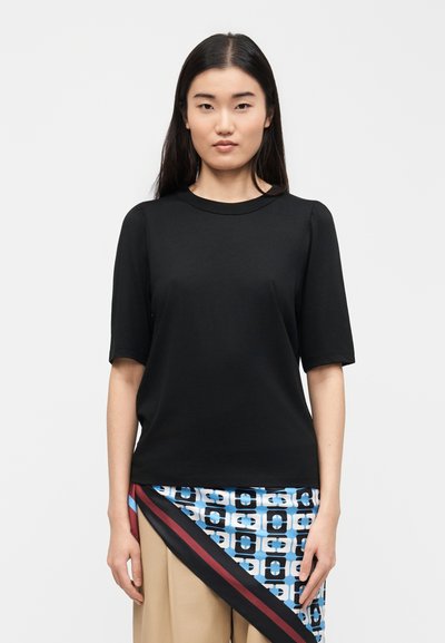 Femme aux cheveux noirs et droits portant un t-shirt noir à manches courtes et un pantalon beige, avec une écharpe enveloppante à motifs bleus sur un côté.
