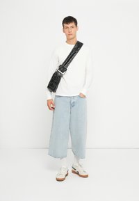 Camicia bianca a maniche lunghe, jeans in denim azzurro chiaro a gamba larga e sneakers bianche con suola marrone. Borsa a tracolla nera con accessori in argento.