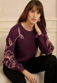 Sweatshirt violet avec broderies florales roses sur les manches, col rond et poignets côtelés. Modèle assis, portant un pantalon sombre.