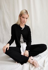 Svart cardigan över en grå skjorta, kombinerad med svarta byxor och rosa skor. Modellen sitter mot en vit bakgrund, med händerna vilande på benen.