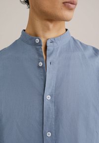 Svetlo modra srajca z gumbki in mandarin ovratnikom, ki ima gladko tkanino, subtilno teksturo in majhne bele gumbke vzdolž sprednje strani.