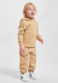 Ein kleines Kind steht, trägt einen beigen Hoodie und passende Jogginghosen, weiße Adidas-Sneaker mit schwarzen Streifen und schaut leicht nach oben.