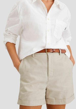 Personne portant un short beige avec les revers retroussés et une ceinture marron, associée à une chemise blanche à boutons avec les manches retroussées.
