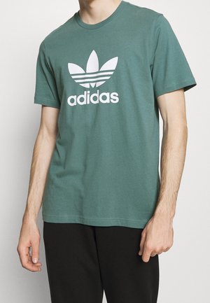 Osoba w zielonej koszulce Adidas z krótkim rękawem z białym logo motywu liścia i czarnych spodniach na jednolitym tle.