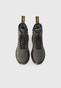 Botas de tornozelo cinzas com uma textura suave, atacadores pretos e forro de pelúcia. Apresenta uma alça com uma marcação amarela e uma sola robusta.