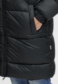 Hand van een persoon in de zak van een zwarte doorgestikte pufferjack met rits en drukknoop, met blauwe jeans aan.