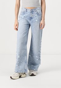 Jeans de mezclilla de pierna ancha en azul claro desgastados con detalles rasgados, cinco bolsillos y cierre de botón. Combinados con zapatillas en gris y crema.