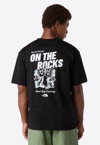 Muž v černém tričku s bílým nápisem "Najdeš mě NA SKALÁCH" a kreslenými kostkami ledu, v kombinaci se světle zelenými kalhotami.