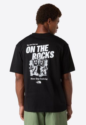 Man wearing black t-shirt with white text "You can find me ON THE ROCKS" and cartoon ice cubes, paired with light green pants.