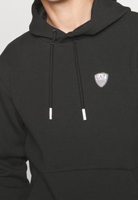 Svart huvtröja i slät tyg med en känguruficka, snörning i huvan med silverfärgade metalldelar och en vit broderad logotyp.