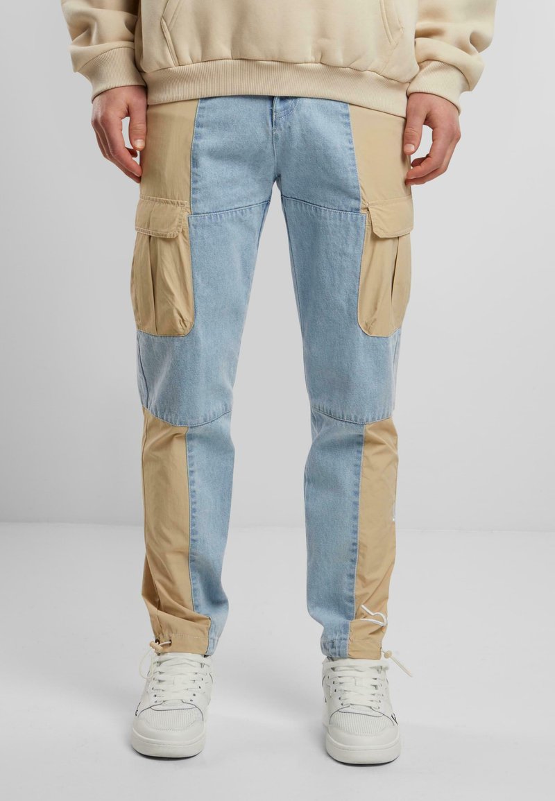 Helle blaue Jeanshose mit beigen Stoffpanelen, Cargo-Taschen und verstellbaren Säumen. Entworfen mit kontrastierenden Texturen und Farben.