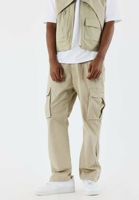 Pantalones cargo beige con múltiples bolsillos laterales, tejido ligero, corte relajado, combinados con una camiseta blanca y zapatillas blancas.