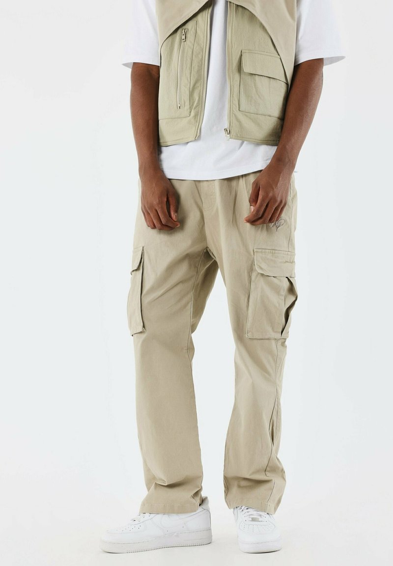 Pantalones cargo beige con múltiples bolsillos laterales, tejido ligero, corte relajado, combinados con una camiseta blanca y zapatillas blancas.