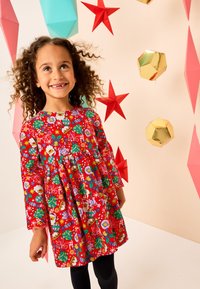 Robe rouge à manches longues avec des motifs festifs comprenant des éléments de Noël, complétée par une taille élastique et un design plissé. Leggings noirs portés.