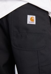 Black work pants made of durable fabric, featuring a patch pocket and a square logo tag with an orange emblem, stitched accents visible.