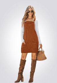Braunes Corduroy-Latzkleid mit Taschen, kombiniert mit einem weißen Langarmoberteil und strukturierten braunen kniehohen Stiefeln. Hält eine braune Handtasche.