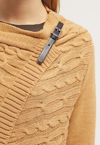 Sweter dziergany z miękkiej, złoto-brązowej przędzy z teksturowanymi wzorami warkoczy. Czarna skórzana klamra zabezpiecza wzór.