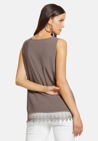 Braunes ärmelloses Top mit einer Spitze am Saum, das eine glatte Textur und eine lockere Passform hat. Getragen mit weißen Hosen.