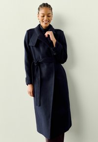 Marineblauwe wollen trenchcoat met een tailleband, brede kraag en zijzakken. De stof heeft een zachte textuur en een klassieke silhouet.