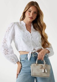 Blusa corta bianca ricamata con maniche lunghe, design floreale e nodo frontale; abbinata a jeans a vita alta blu chiaro e una piccola borsa beige.