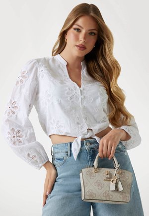 Blusa corta bianca ricamata con maniche lunghe, design floreale e nodo frontale; abbinata a jeans a vita alta blu chiaro e una piccola borsa beige.