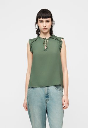 Ung kvinde iført grøn ærmeløs bluse med flæsekant ved skuldrene og bindebånd ved halsen, kombineret med lyseblå jeans, stående mod en hvid baggrund.