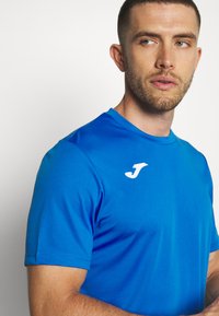Modré sportovní tričko s krátkými rukávy. Vyrobeno z hladké tkaniny, s bílým logem v levém horním rohu a jednoduchým, čistým designem.