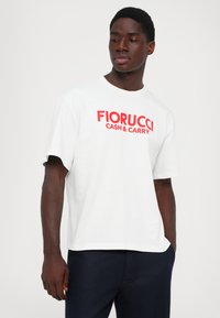Jauns vīrietis valkā baltu T-kreklu ar sarkanu tekstu "FIORUCCI CASH & CARRY", stāv ar vienu roku tumšzilos biksēs, skatoties pa šķelmi pa kreisi.