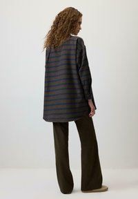 Majica s pregotkami rokavi z različnimi modrimi in rjavimi črtami, kombinirana z širokimi hlačami v olivno zeleni barvi. Mehka tekstura, vidni gumbi na manšetah.