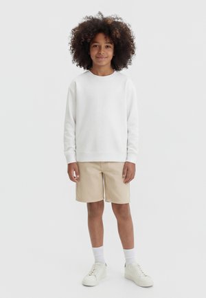 Lokkis juustega laps, kes kannab valget pikade varrukatega särki, beeže pükse, valgeid sokke ja valgeid tenniseid, seisab ühevärvilise tausta ees.