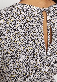 Mustakankainen kangas, jossa on valkoisia ja keltaisia kukkakuoseja; sisältää avainaukkojatkeen, jossa on rusetti. Pinta näyttää sileältä.