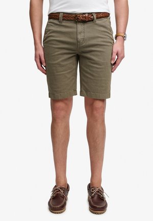 Mann trägt olivgrüne Shorts mit braunem geflochtenem Gürtel, braune Leder-Bootsschuhe, ein weißes Hemd und eine beige Uhr am linken Handgelenk.