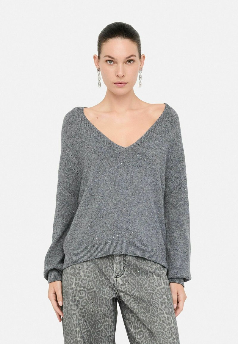 Gray knitted sweater with a deep V-neck, loose-fit design, and ribbed cuffs. Paired with patterned gray jeans featuring a subtle texture.