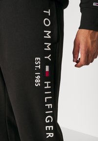 Černé tepláky s bílým vyšívaným textem "TOMMY HILFIGER" a "EST. 1985", se známkou malého barevného loga na boku.