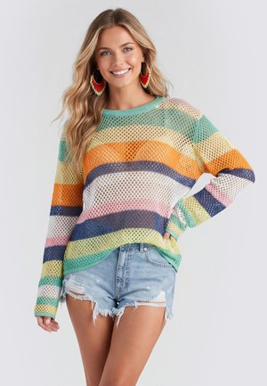 Lächelnde Frau mit langen blonden Haaren, die große bunte Ohrringe, einen mehrfarbigen Netzpullover und zerrissene Jeansshorts trägt und vor einem hellen Hintergrund posiert.